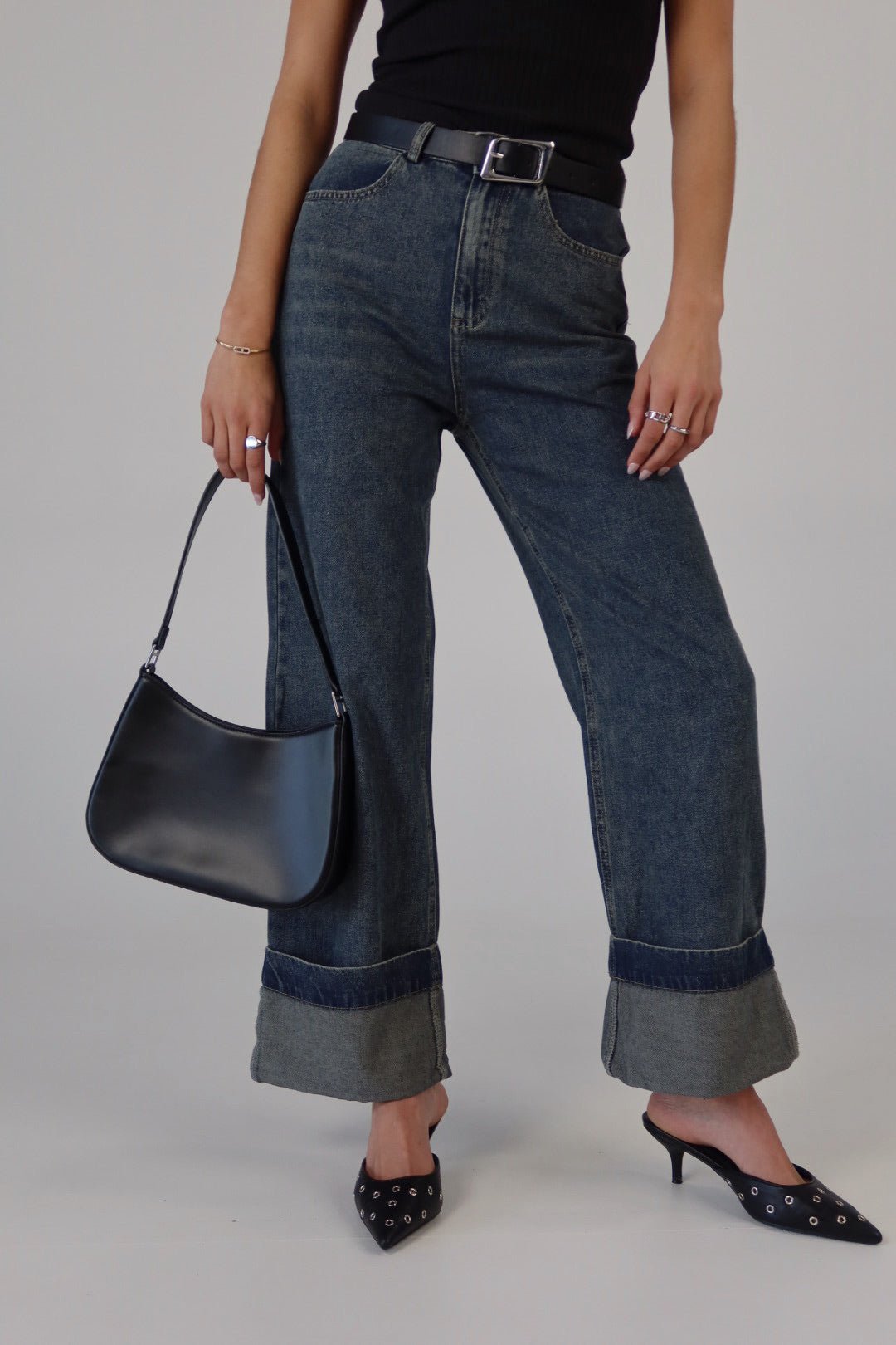 A person standing wearing fold-over washed denim pants with a black belt, black bag, and black shoes.