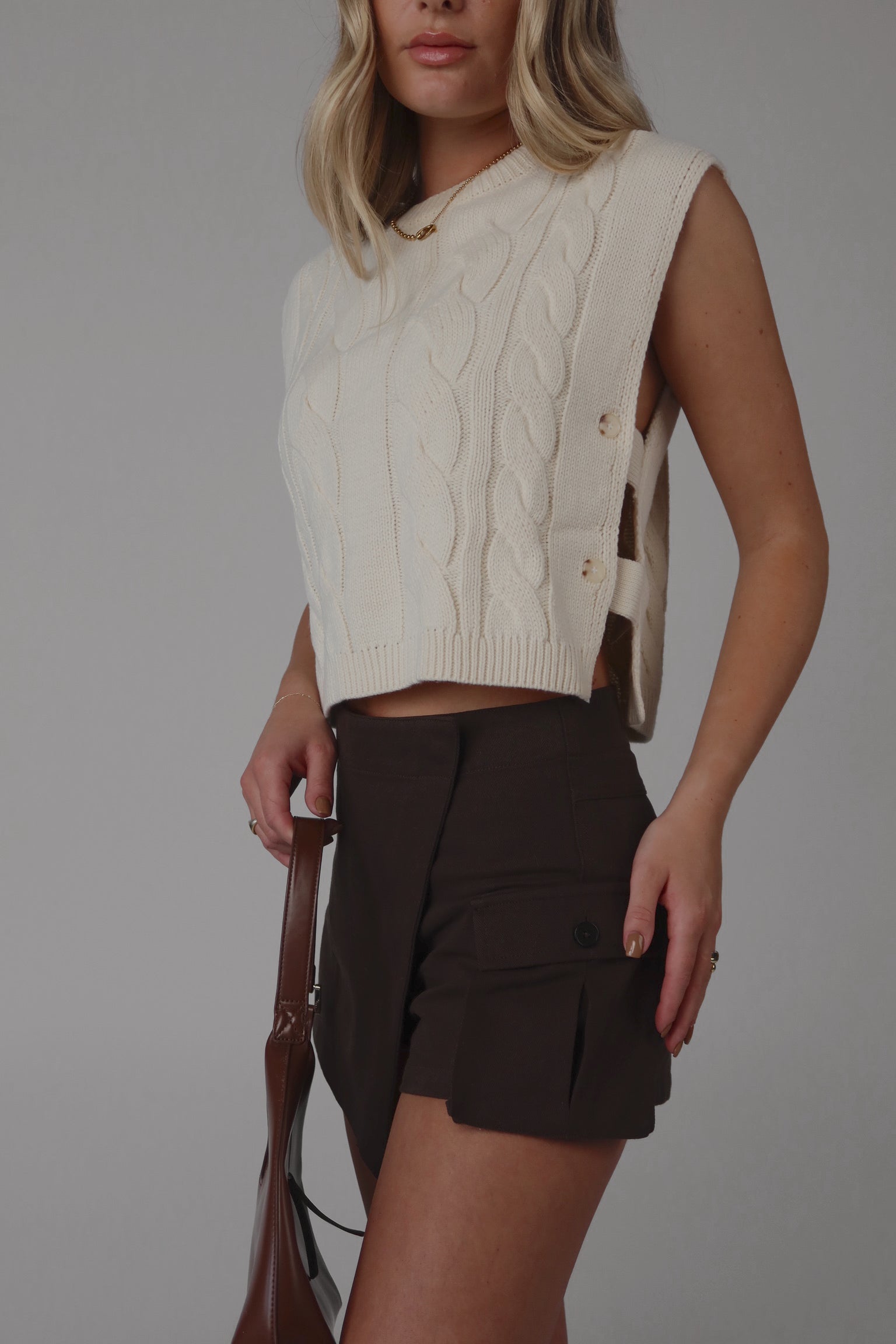A female model wearing a beige sleeveless cable knit sweater with an open side design and button closure, paired with brown shorts.